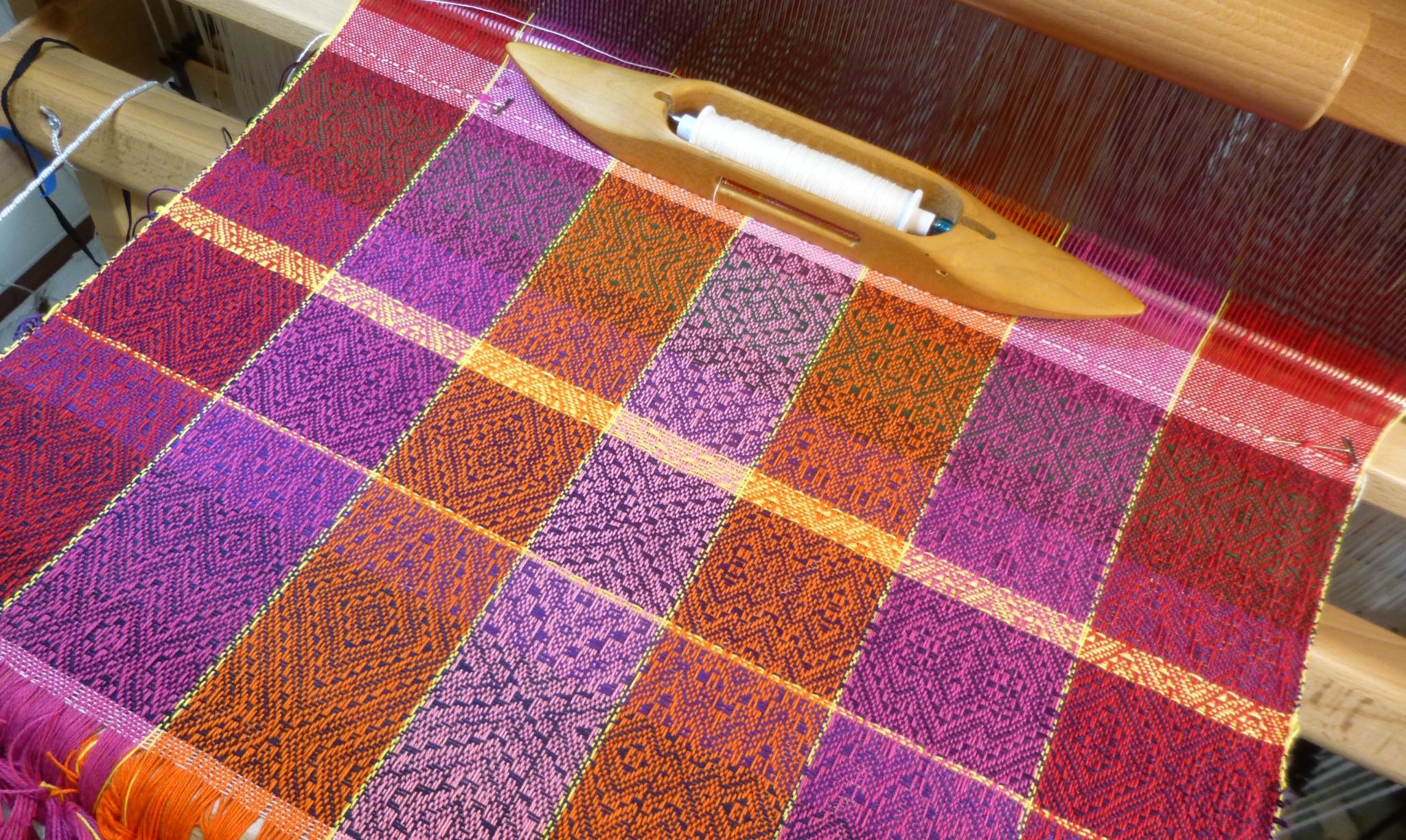 A project gamp in pink, orange, purple and red on the loom, blocks of different twills separated by yellow lines, with a boat shuttle resting on the cloth. Woven by Roxie Rochat.